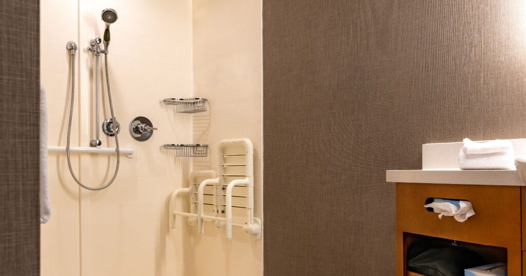 Accessible walk-in shower with grab bars, handheld showerhead, and fold-down seat next to a modern vanity.
